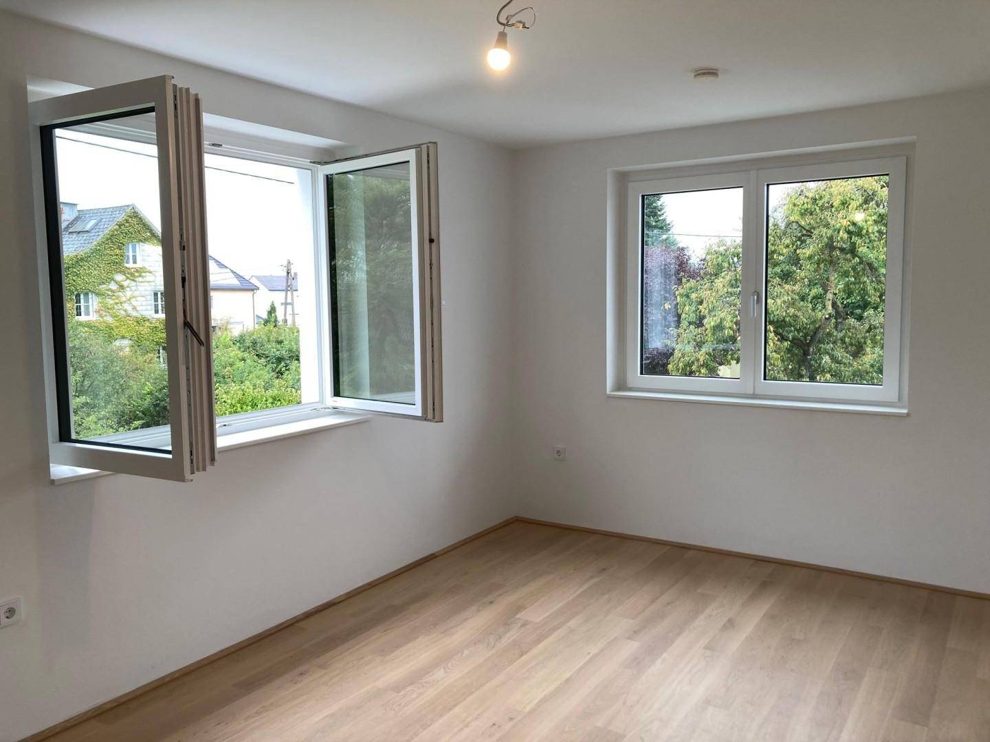 Leerer Raum mit Holzboden, weißen Wänden und zwei Fenstern mit Blick ins Grüne.