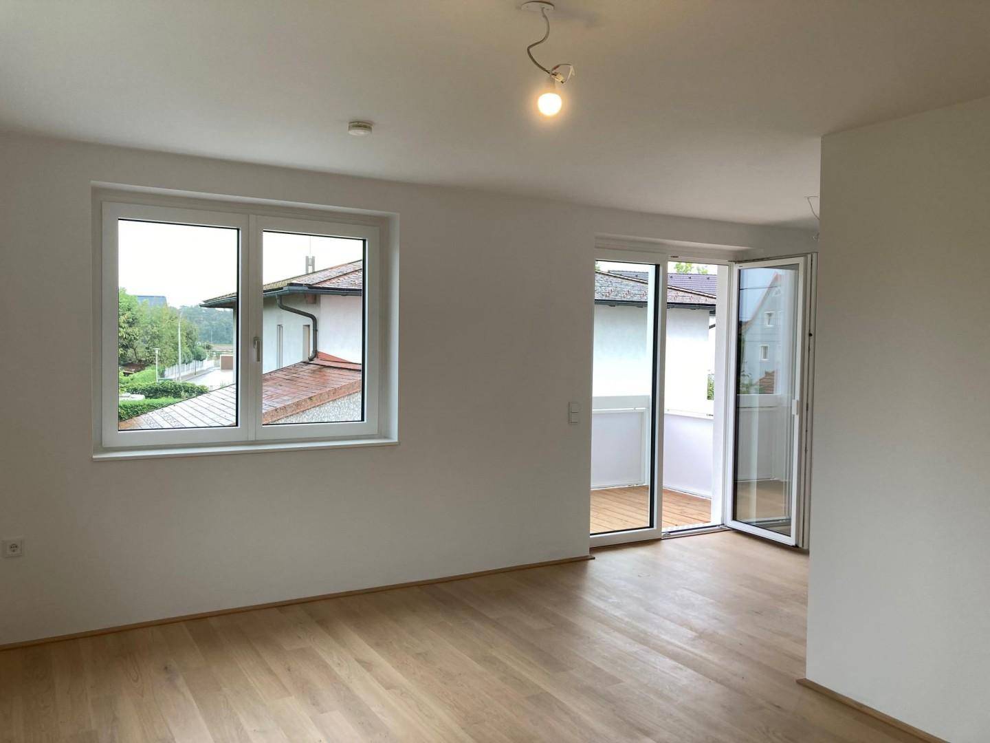 Heller, leerer Raum mit Holzboden, großen Fenstern und Zugang zu einem Balkon.