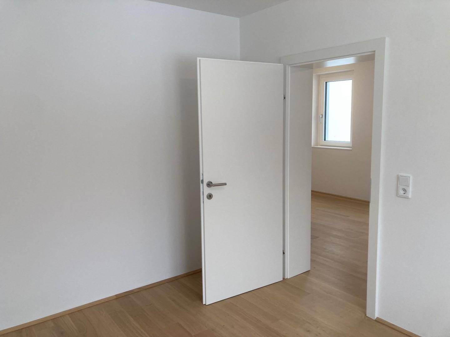 Leerer Raum mit weißer Tür, die den Blick in einen weiteren hellen Raum freigibt.