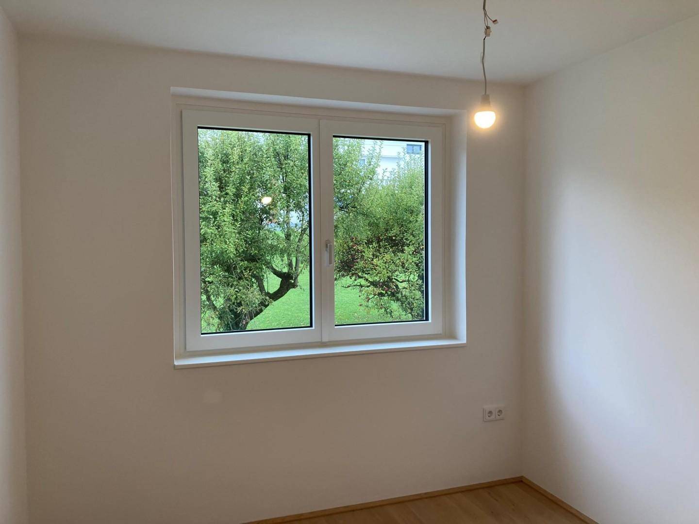 Leerer Raum mit hellem Holzfußboden und Fenster mit Blick ins Grüne.
