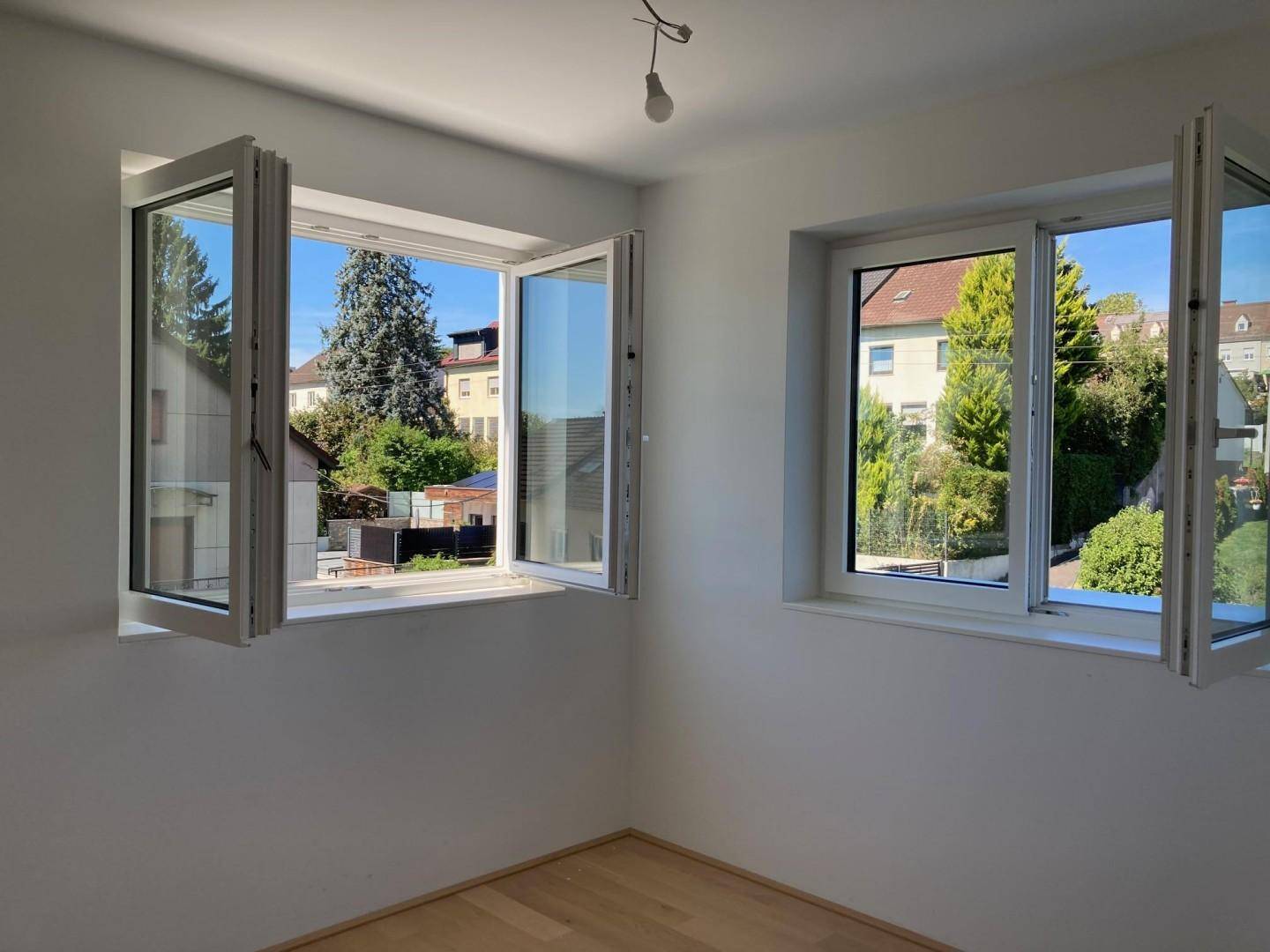 Heller Raum mit zwei großen, geöffneten Fenstern, die einen Blick ins Grüne und auf Nachbarhäuser bieten.