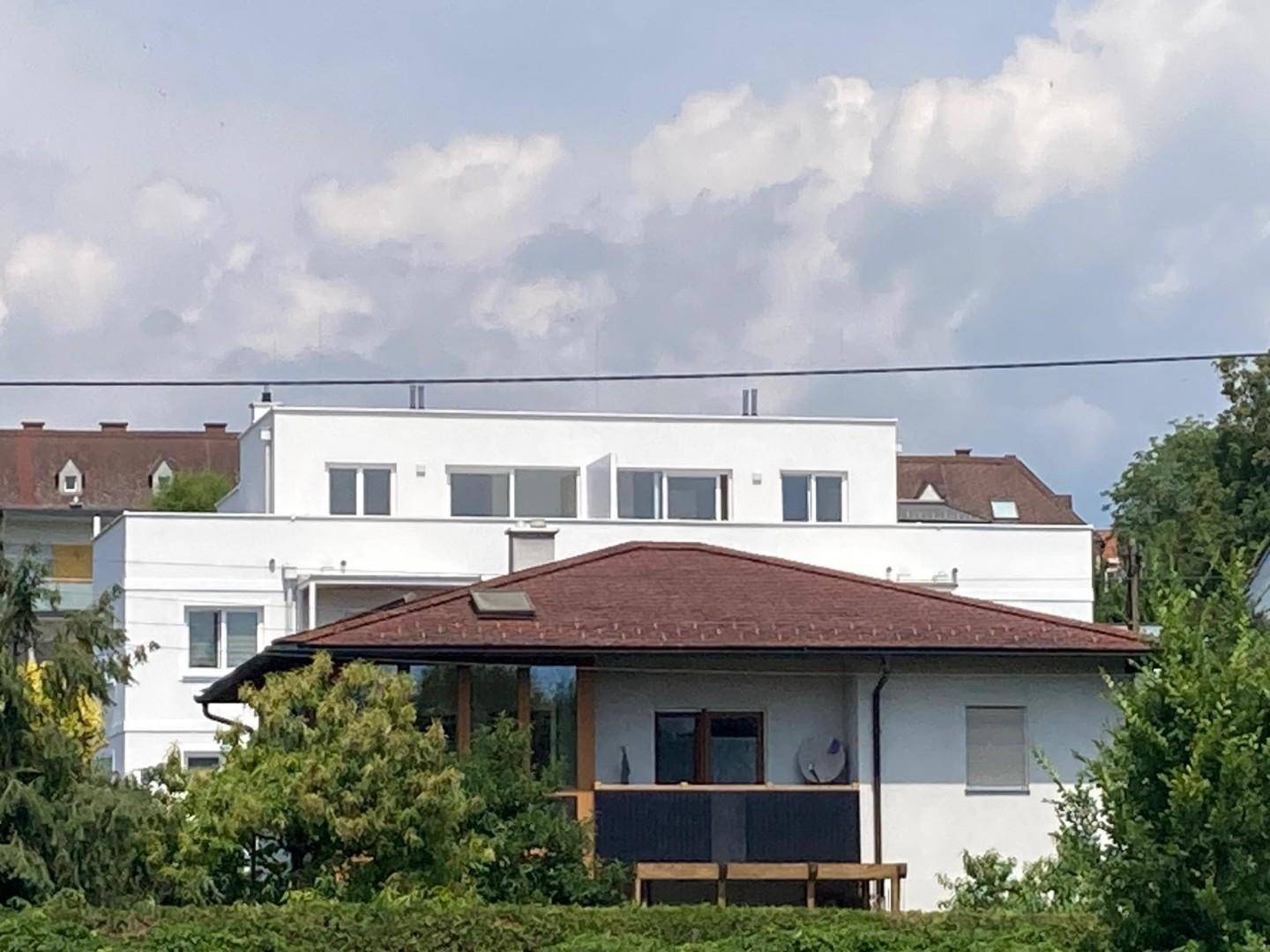 Weißes Mehrfamilienhaus mit Flachdach und großen Fenstern, umgeben von Bäumen unter bewölktem Himmel.