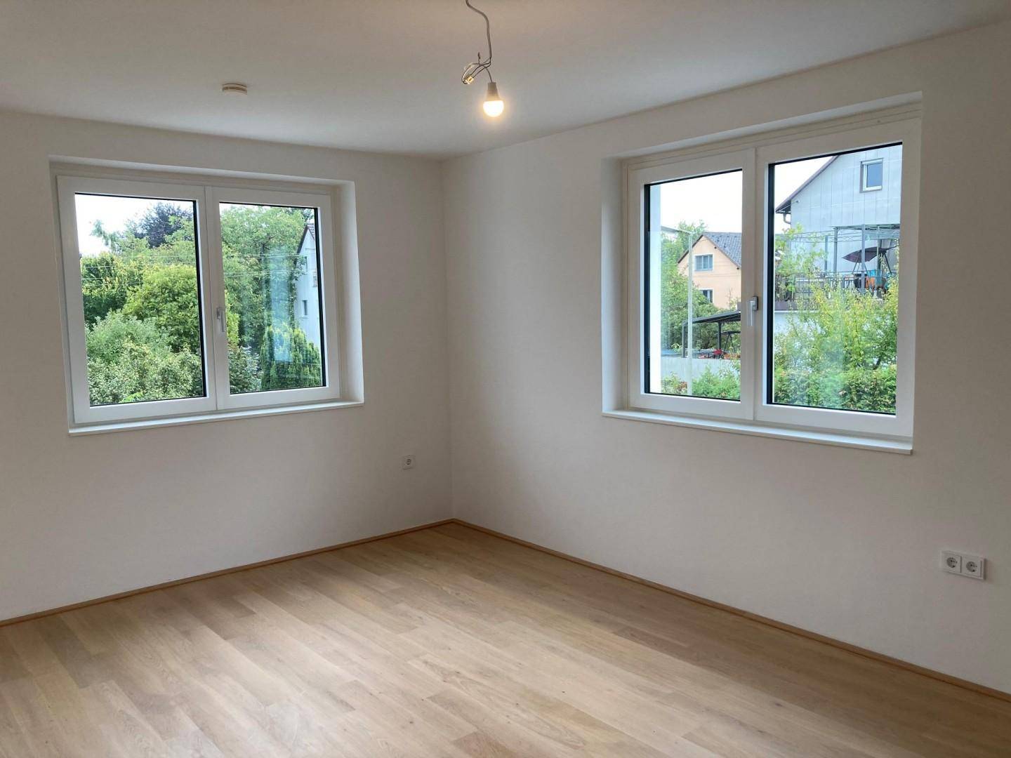 Geräumiges Zimmer mit Holzboden und zwei großen Fenstern, die einen Blick ins Grüne bieten.