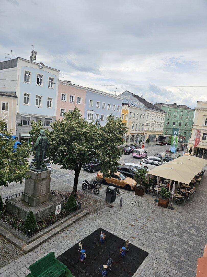Blick von oben auf einen belebten Stadtplatz mit historischen Gebäuden, Bäumen und Außengastronomie.