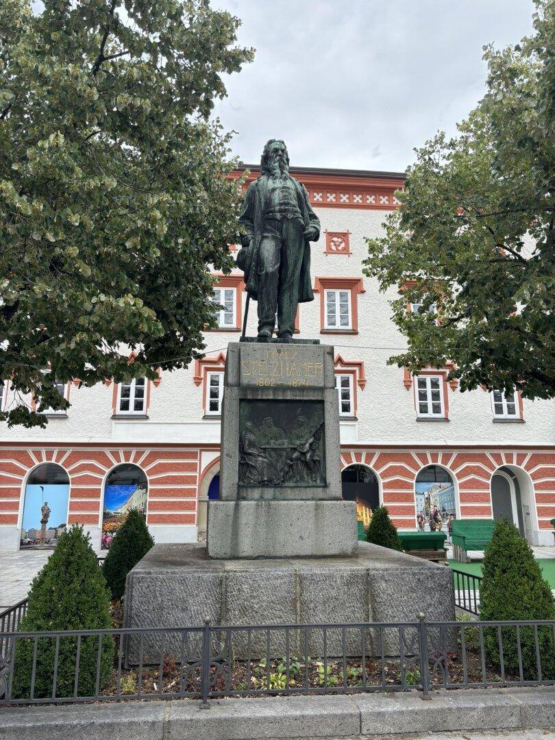 Denkmal auf einem Stadtplatz mit dem historischen Gebäude im Hintergrund und Bäumen.