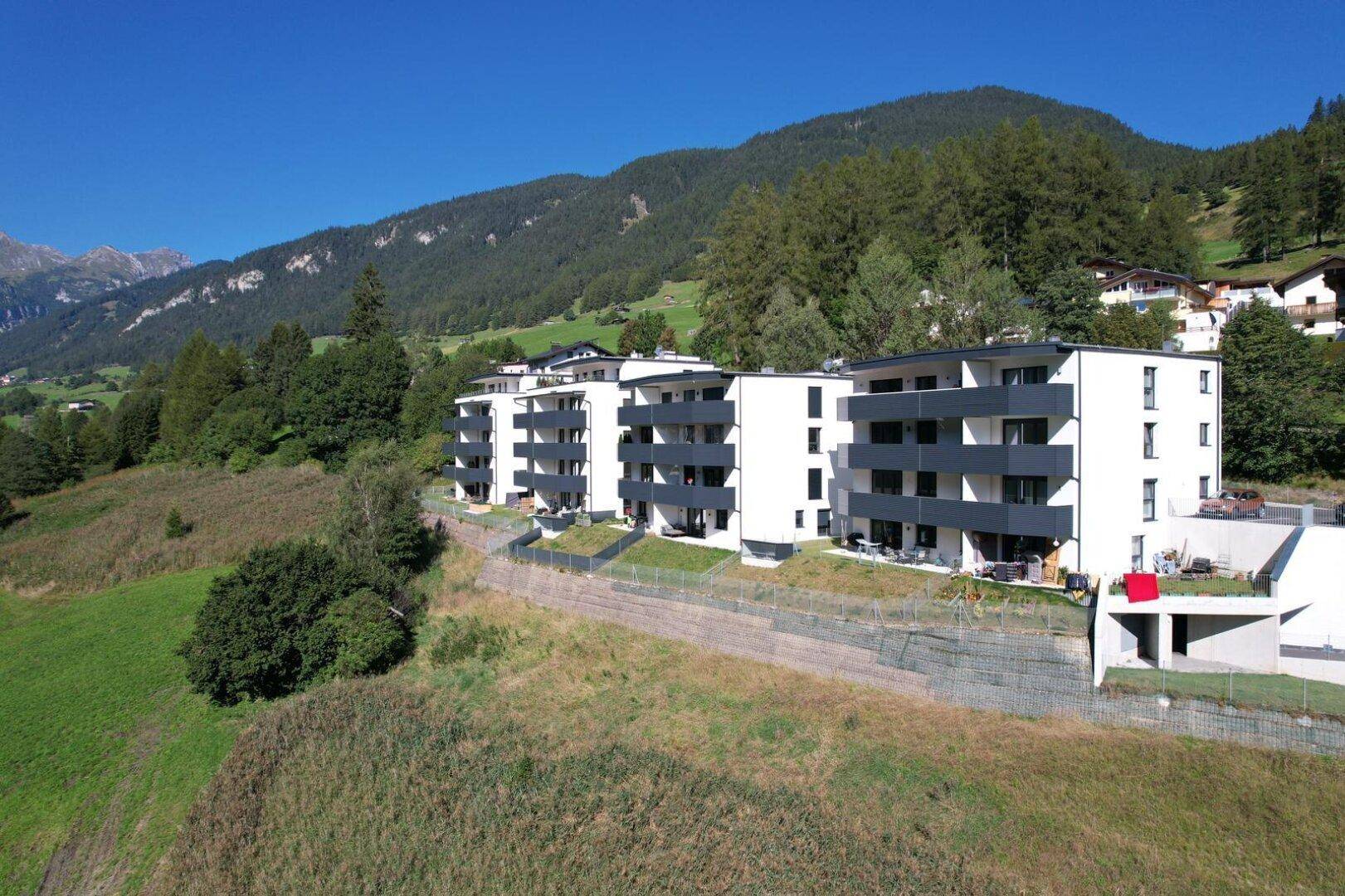 Neue Wohngebäude mit weißer Fassade und dunklen Balkonen, umgeben von grünen Hängen und Bergen.