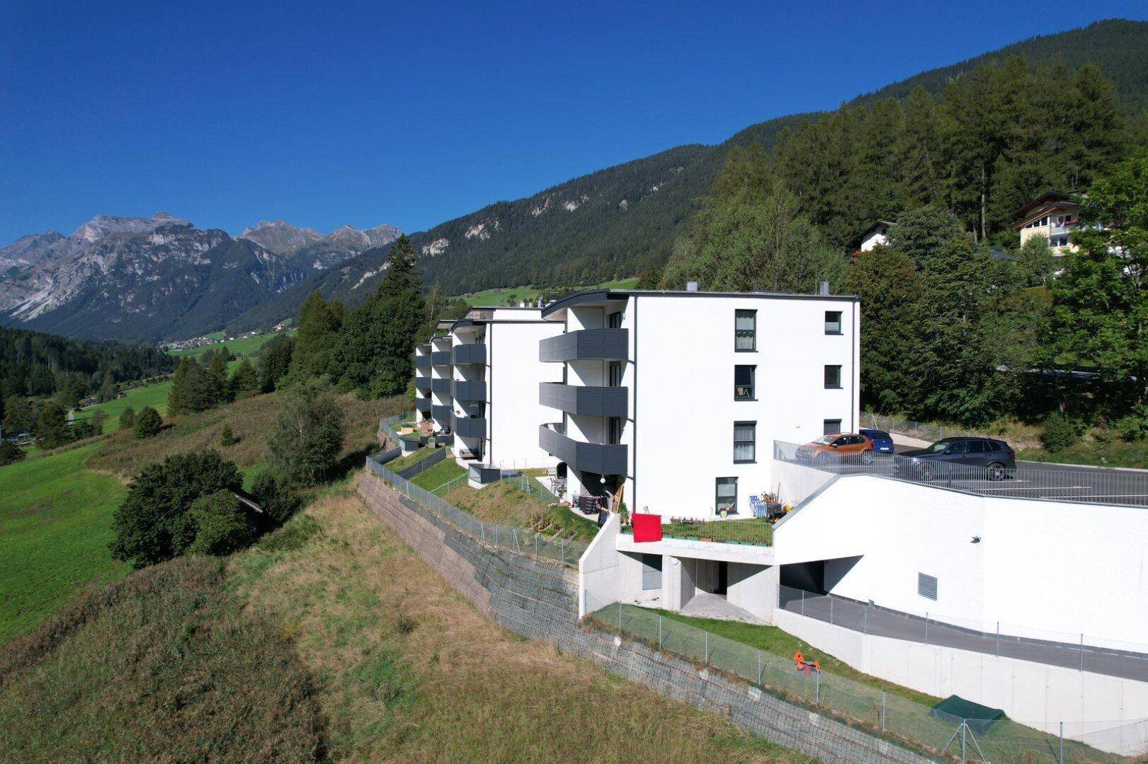Moderne Wohngebäude mit Tiefgaragenzufahrt und kleinen Außenbereichen in alpiner Umgebung.