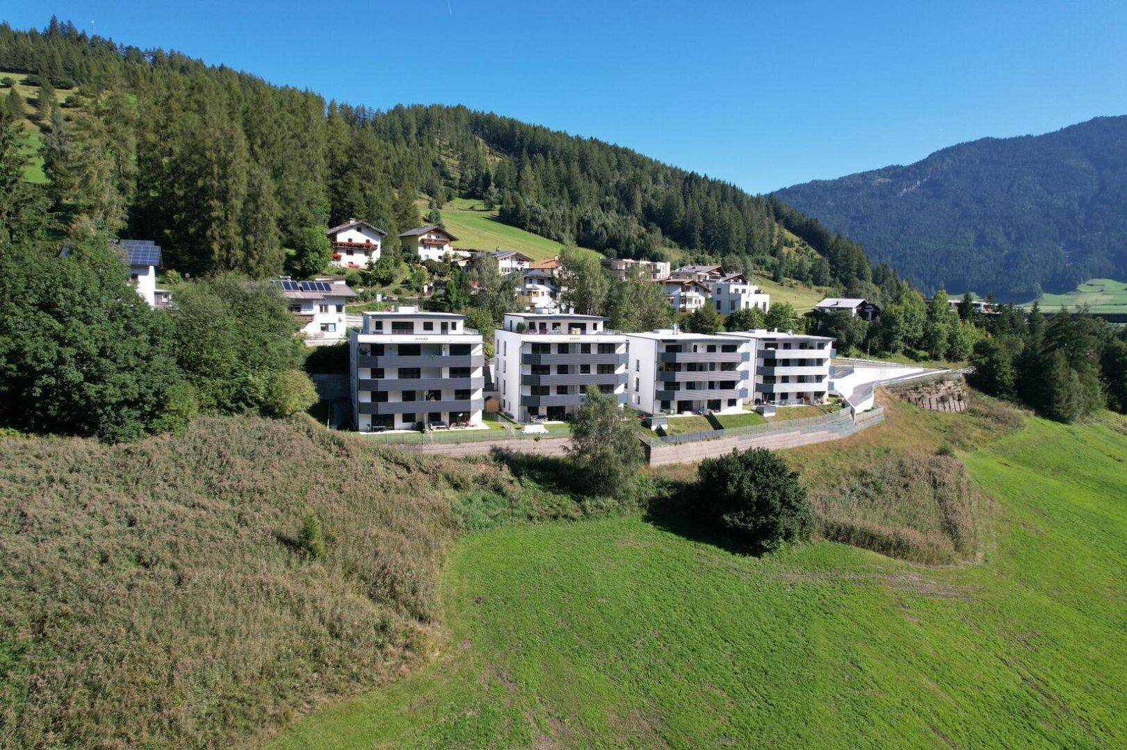 Moderne Wohngebäude auf einem Hügel, umgeben von grünen Wiesen und Wäldern mit Bergblick.