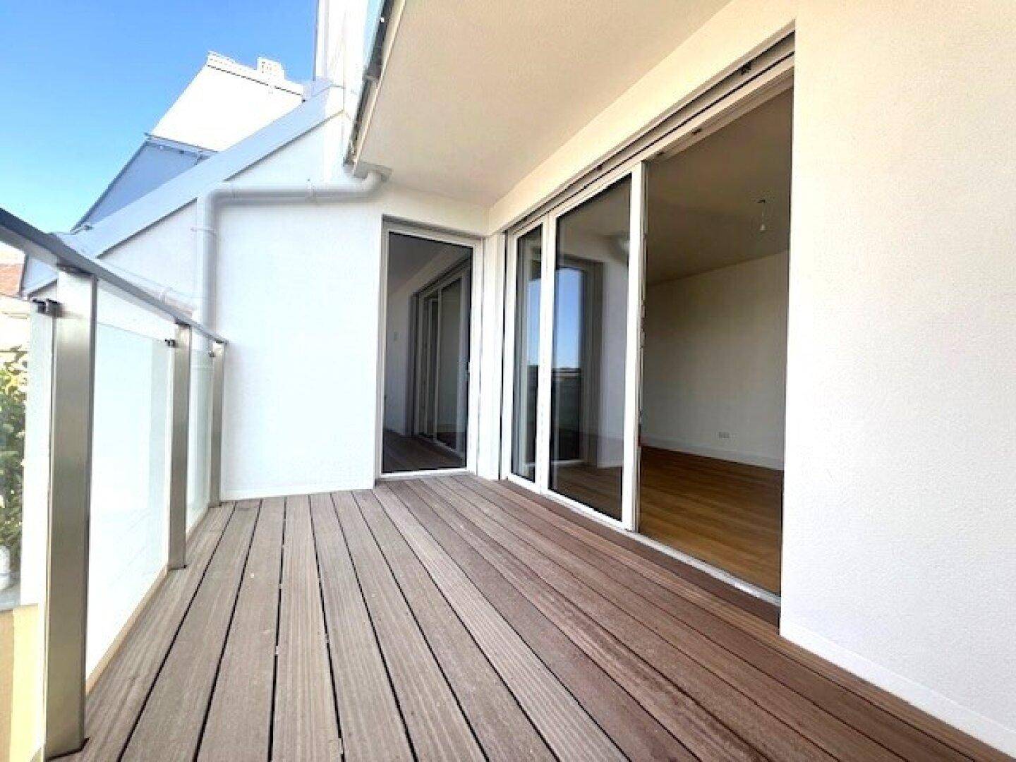 Geräumiger Balkon mit Holzboden und Glasgeländer, der Zugang zu hellen Innenräumen bietet.
