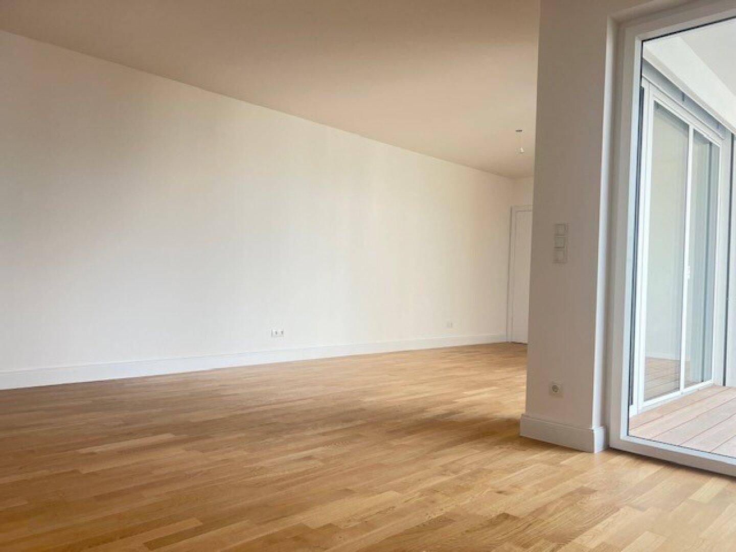 Geräumiger, unmöblierter Raum mit hellem Parkettboden und Zugang zu einem Außenbereich.