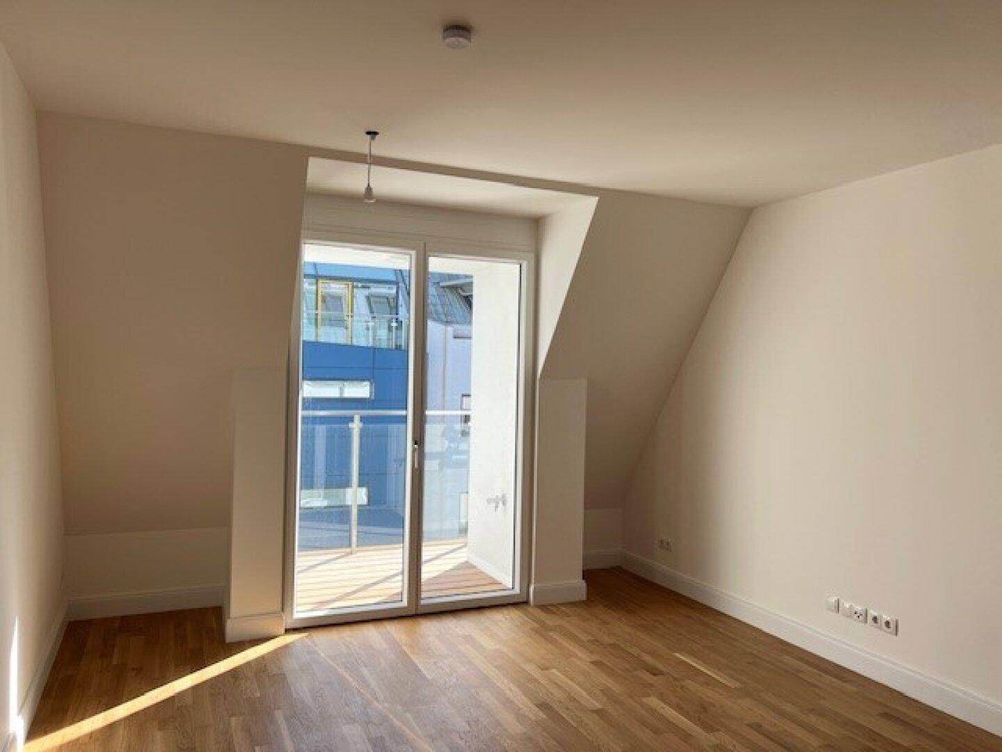 Leerer Raum mit Holzboden, schräger Decke und großem Fenster mit Zugang zum Balkon.