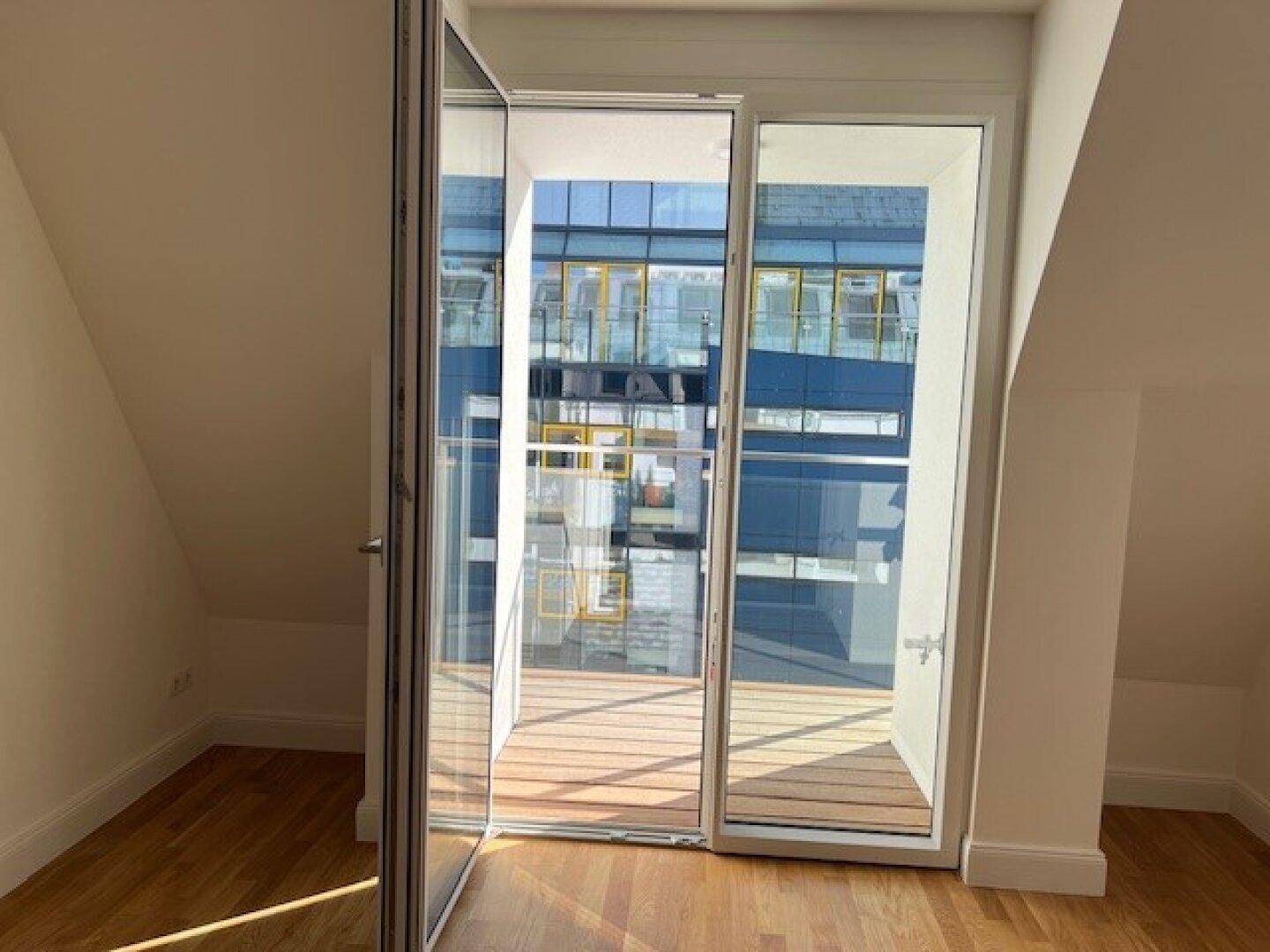 Heller Innenraum mit Holzboden und Zugang zu einem sonnigen Balkon, mit schräger Decke.