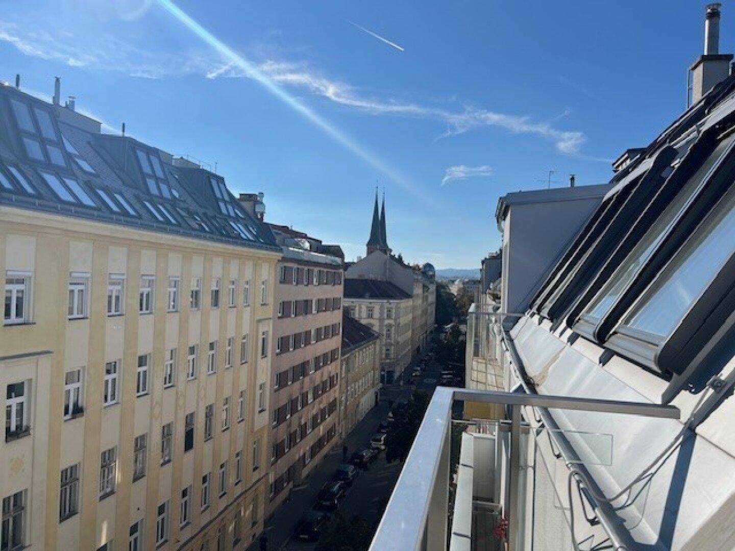 Sonniger Blick vom Balkon über eine belebte Stadtstraße mit historischen Gebäuden und Kirchturm.