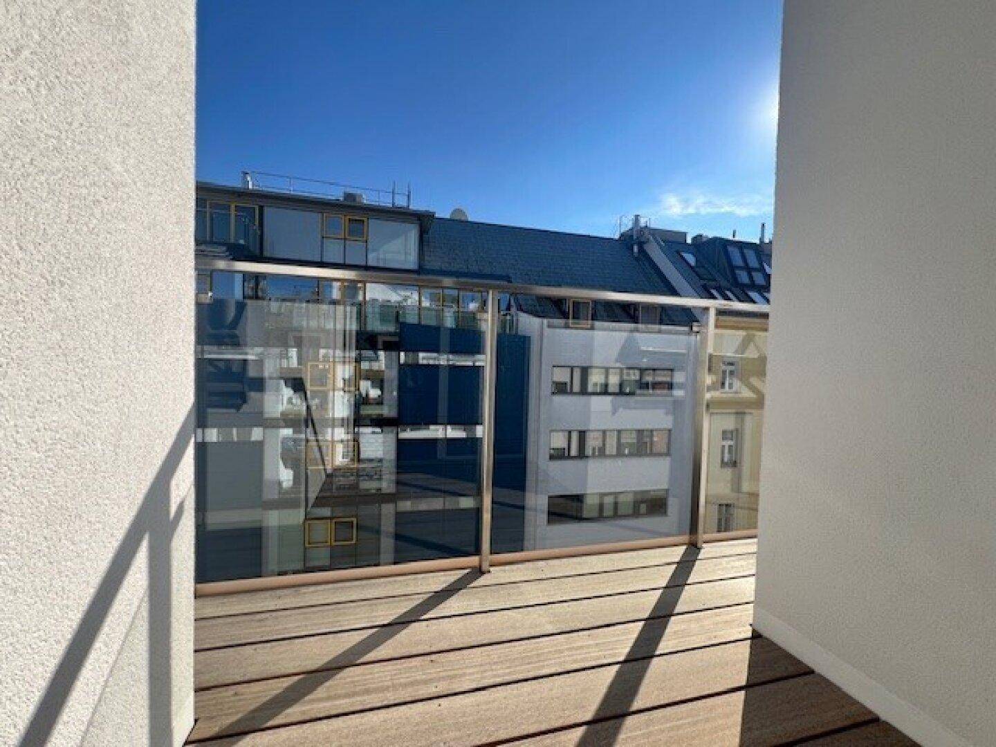 Heller Balkon mit Holzboden und moderner Glasbrüstung, bietet Blick auf gegenüberliegende Gebäude.