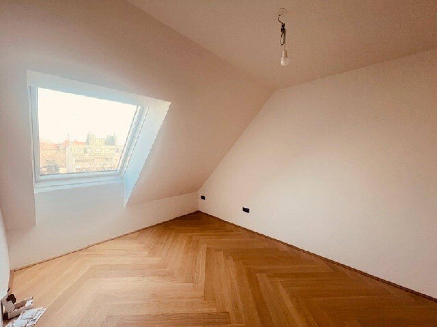Helles Dachzimmer mit schrägen Wänden, Fischgrätparkett und einem großen Dachfenster.