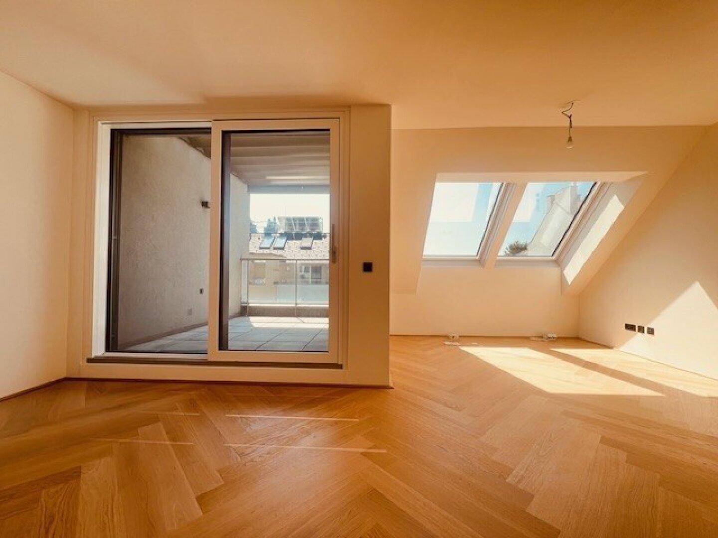 Lichtdurchfluteter Raum mit Fischgrätparkett, Zugang zum Balkon und schrägen Wänden im Dachgeschoss.