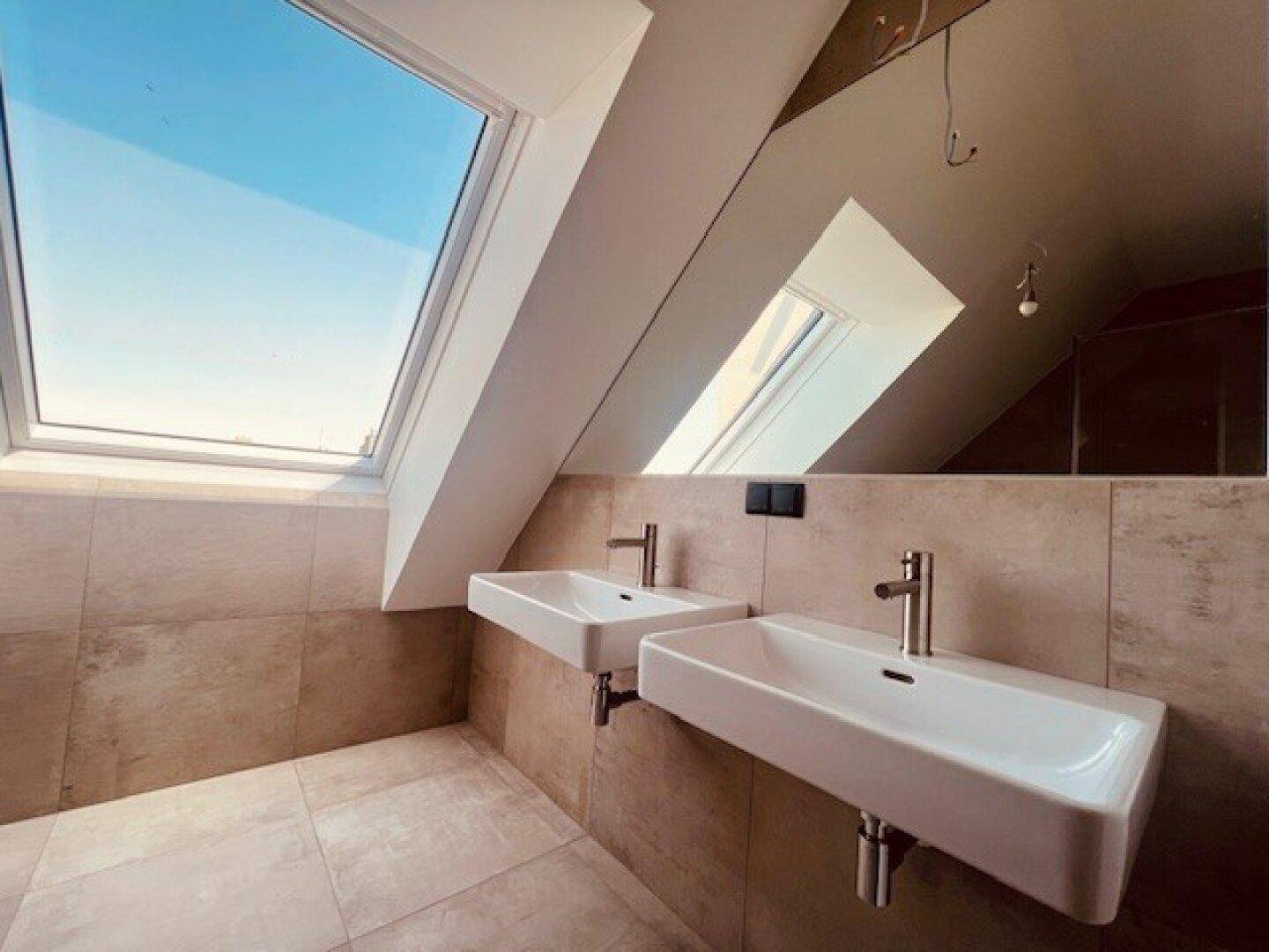 Modernes Badezimmer mit zwei Waschbecken, beigefarbenen Fliesen und einem großen Dachfenster.