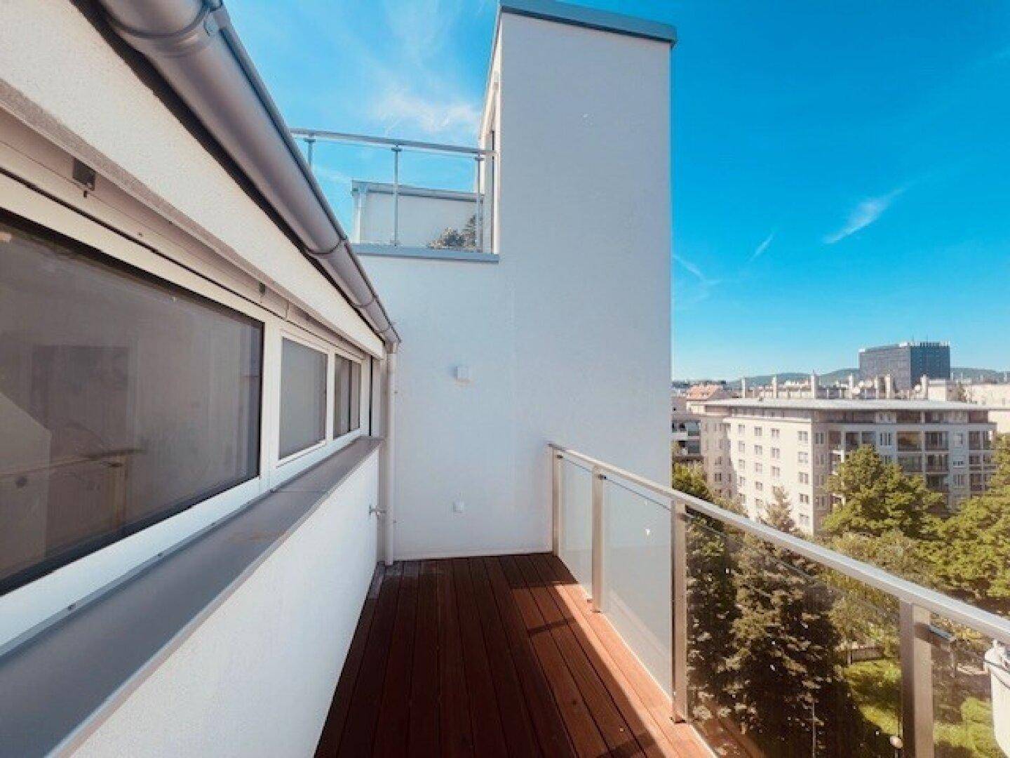 Schmaler Balkon mit Holzboden und Glasgeländer, bietet einen Blick auf Bäume und umliegende Gebäude.