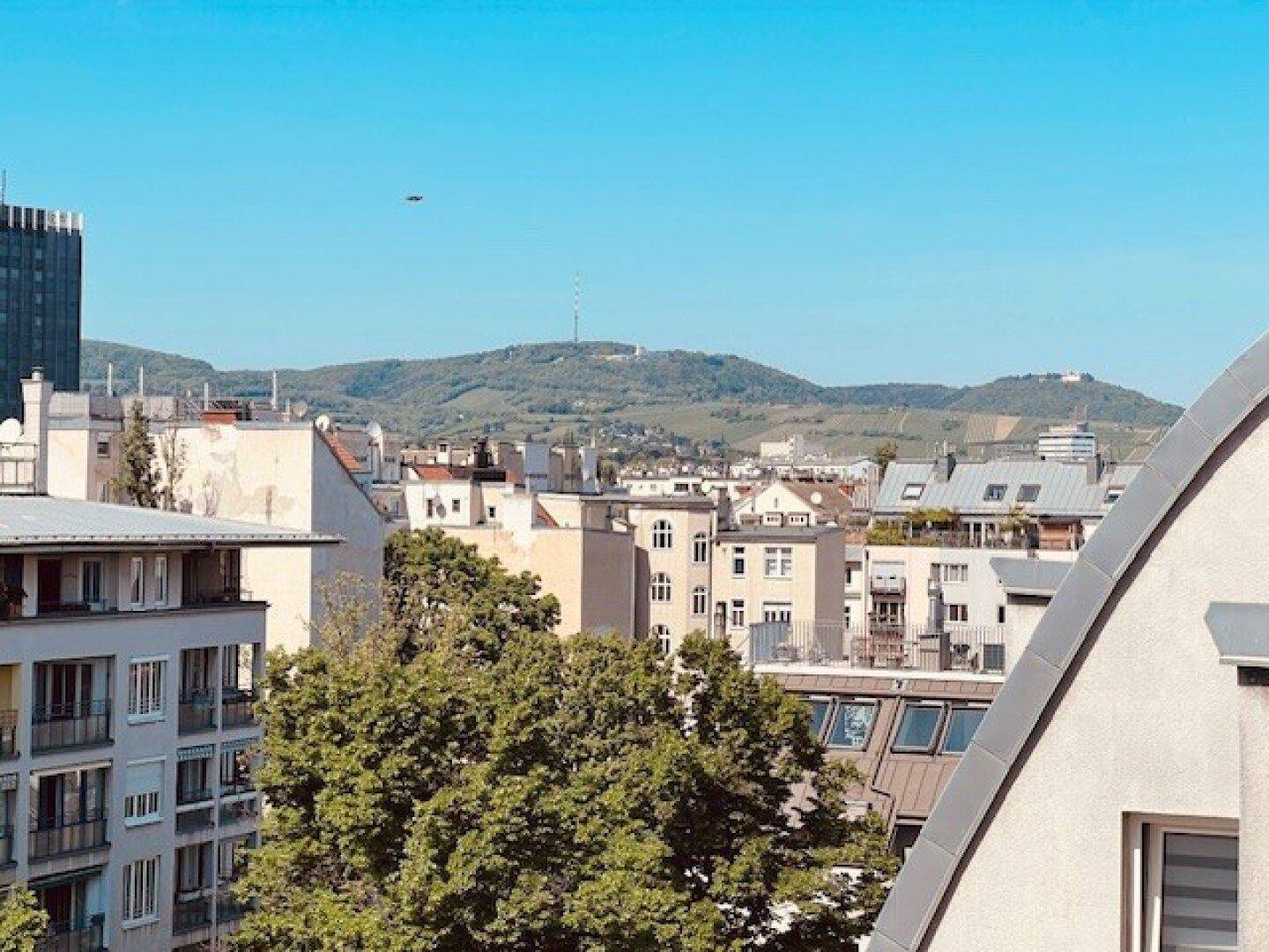 Panoramablick über die Stadt mit grünen Hügeln und Bergen im Hintergrund unter klarem Himmel.