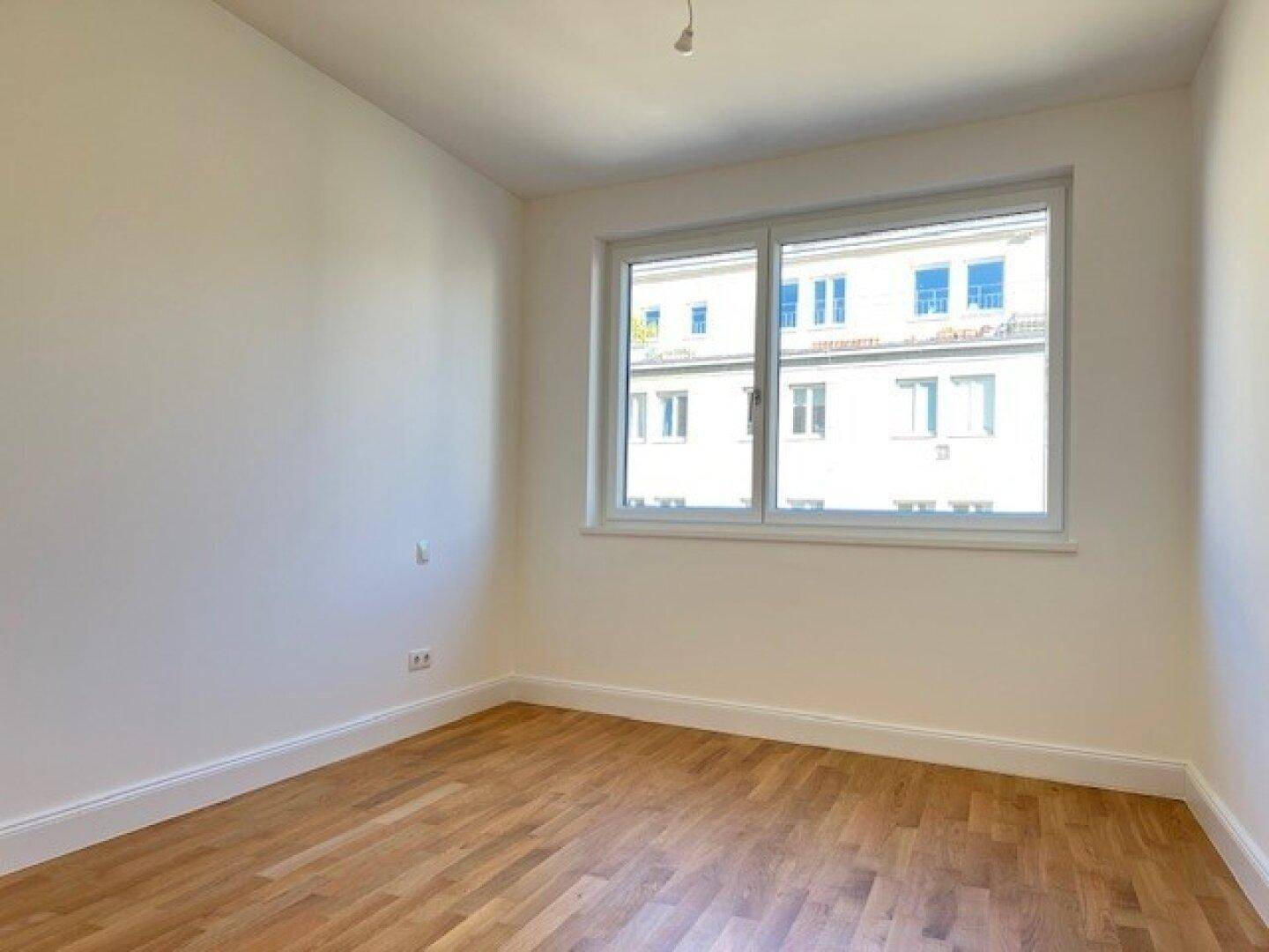 Leeres Zimmer mit Holzboden und großem Fenster, das viel Tageslicht hereinlässt.