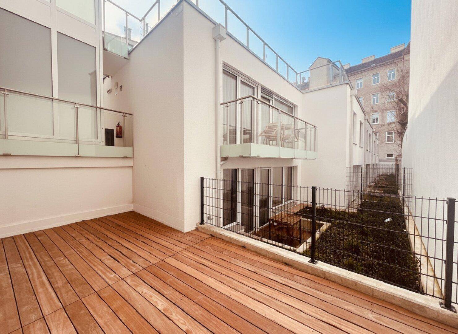 Geräumige, unmöblierte Terrasse mit Holzdeck und Blick auf die umliegenden Gebäude und einen kleinen Gartenbereich.