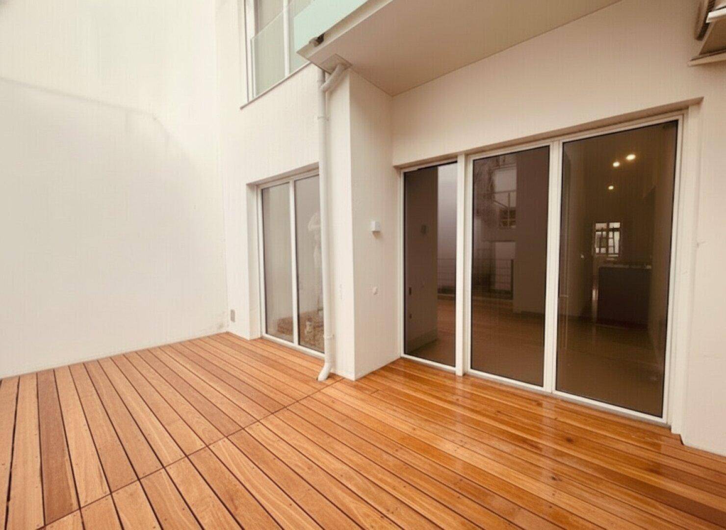 Leere Terrasse mit hellem Holzdeck und großen Glasschiebetüren, bereit für individuelle Gestaltung und Nutzung.