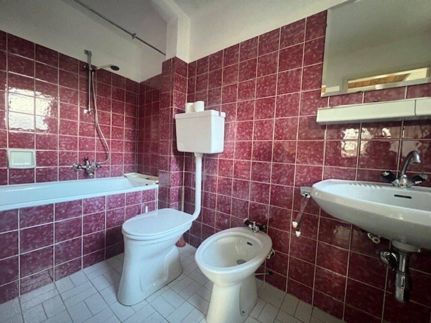 Badezimmer im Originalzustand mit roten Fliesen, Badewanne, Toilette und Bidet.