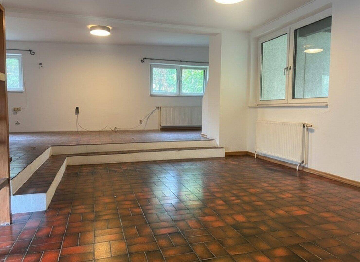 Geräumiger, unmöblierter Wohnbereich mit gefliestem Boden und Fenstern, bereit zur Renovierung.