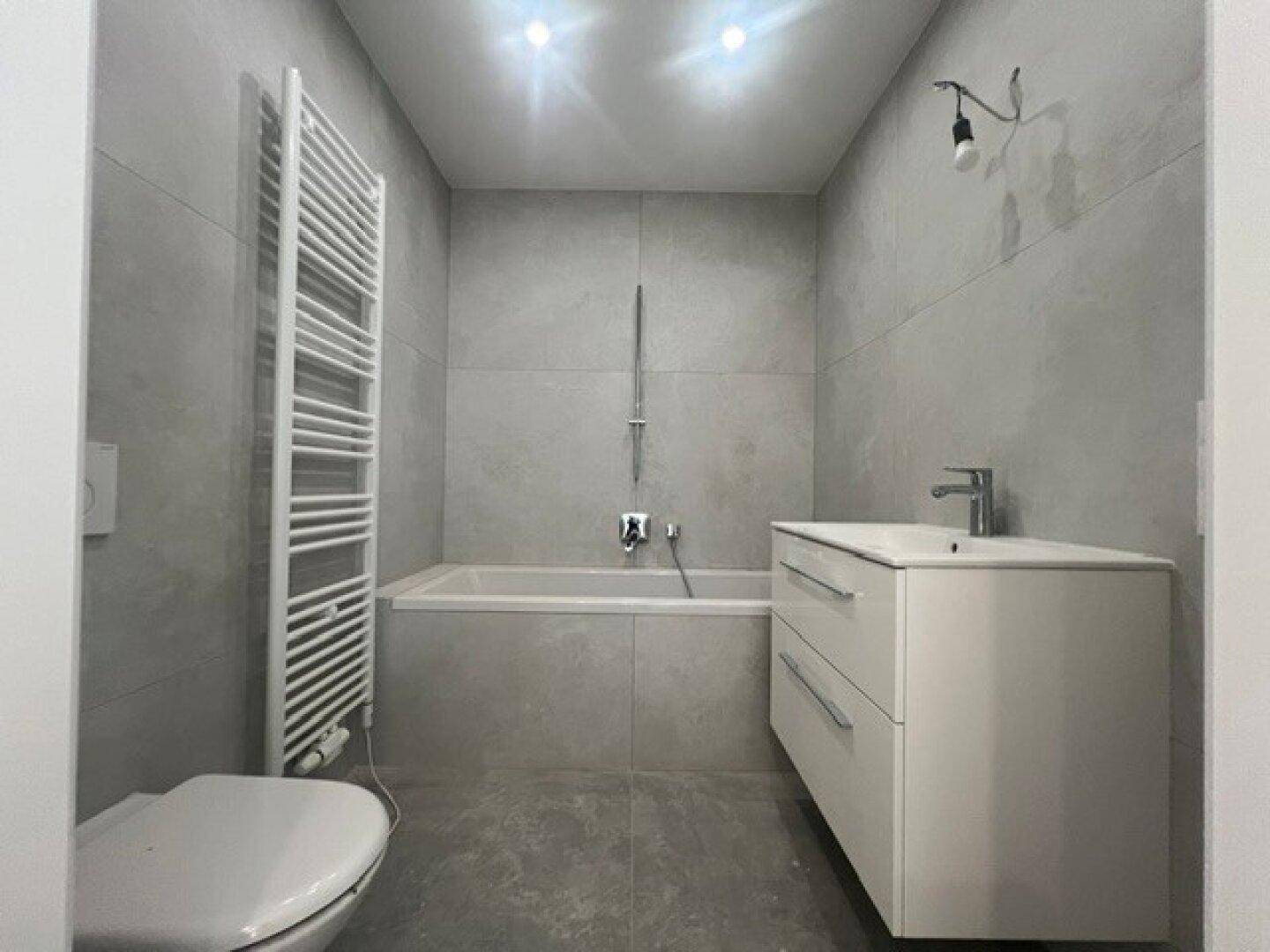 Modernes Badezimmer mit grauen Fliesen, Badewanne, wandhängendem WC und Handtuchheizkörper.