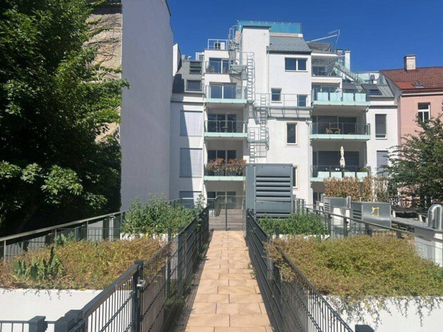 Modernes Apartmentgebäude mit mehreren Balkonen und einem begrünten Dachgartenweg unter blauem Himmel.