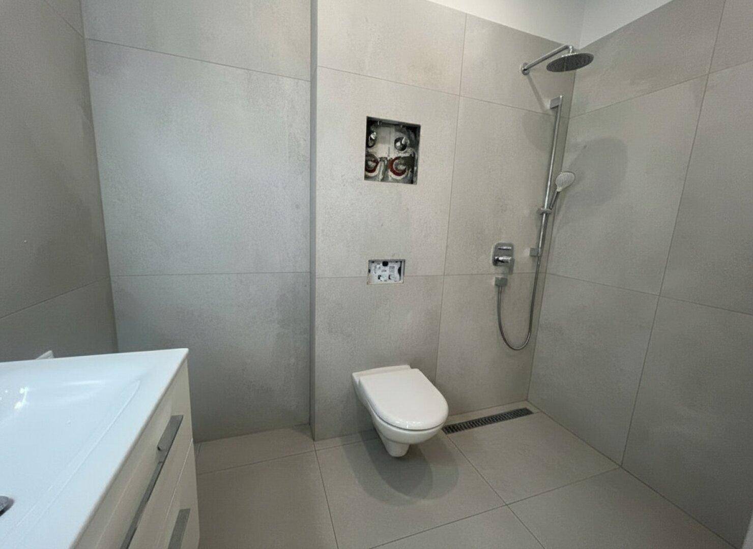 Modernes Badezimmer mit grauen Fliesen, wandhängendem WC und einer geräumigen Duschzone.