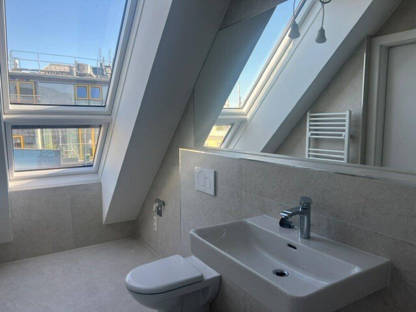 Modernes Badezimmer im Dachgeschoss mit Waschbecken, Toilette, beheiztem Handtuchhalter und Dachfenstern.
