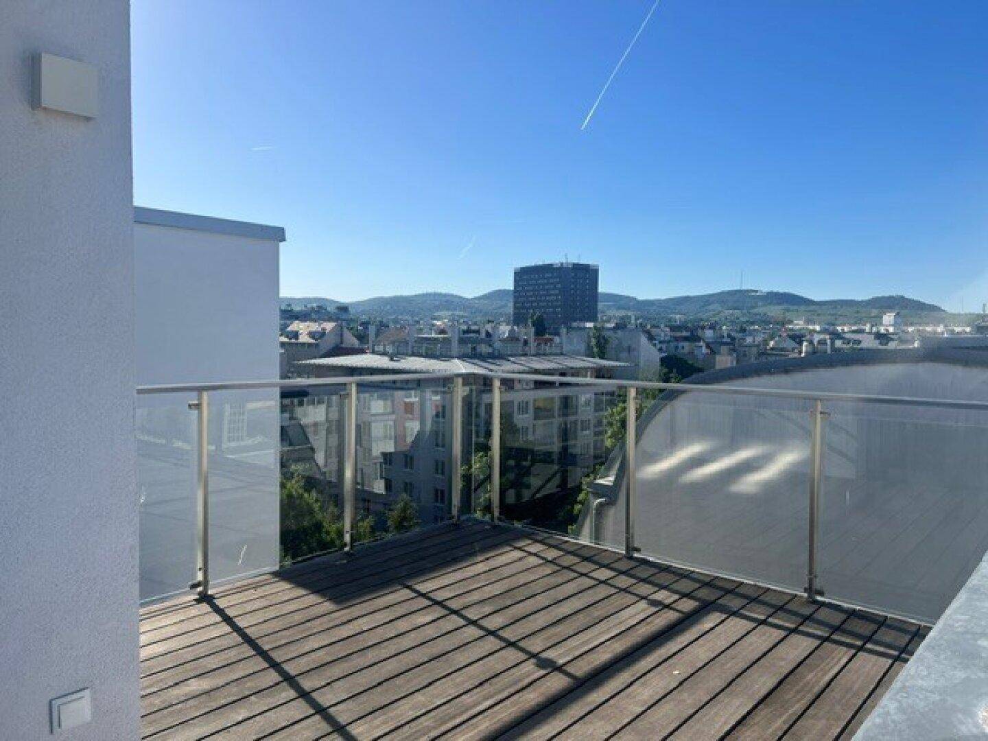 Dachterrasse mit Glasgeländer und Holzboden, bietet einen Panoramablick über die Stadt und die Berge.