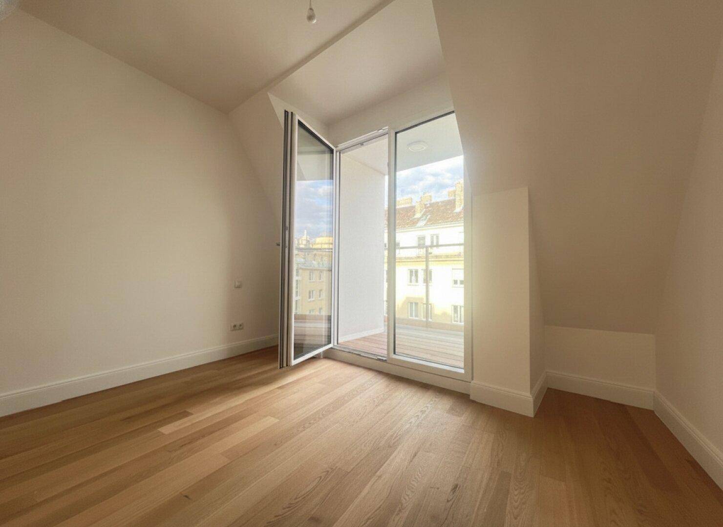 Heller Raum mit Holzboden, Dachschräge und einer Fenstertür zu einem kleinen Balkon.
