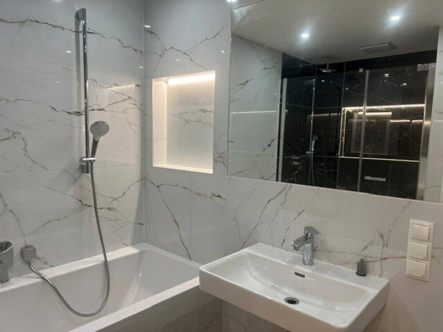 Helles Badezimmer mit Marmorfliesen, Badewanne, Einzelwaschbecken und beleuchteter Nische.