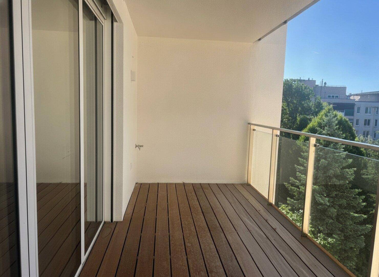 Geräumiger Balkon mit Holzbelag und Glasgeländer, bietet einen sonnigen Blick ins Grüne.