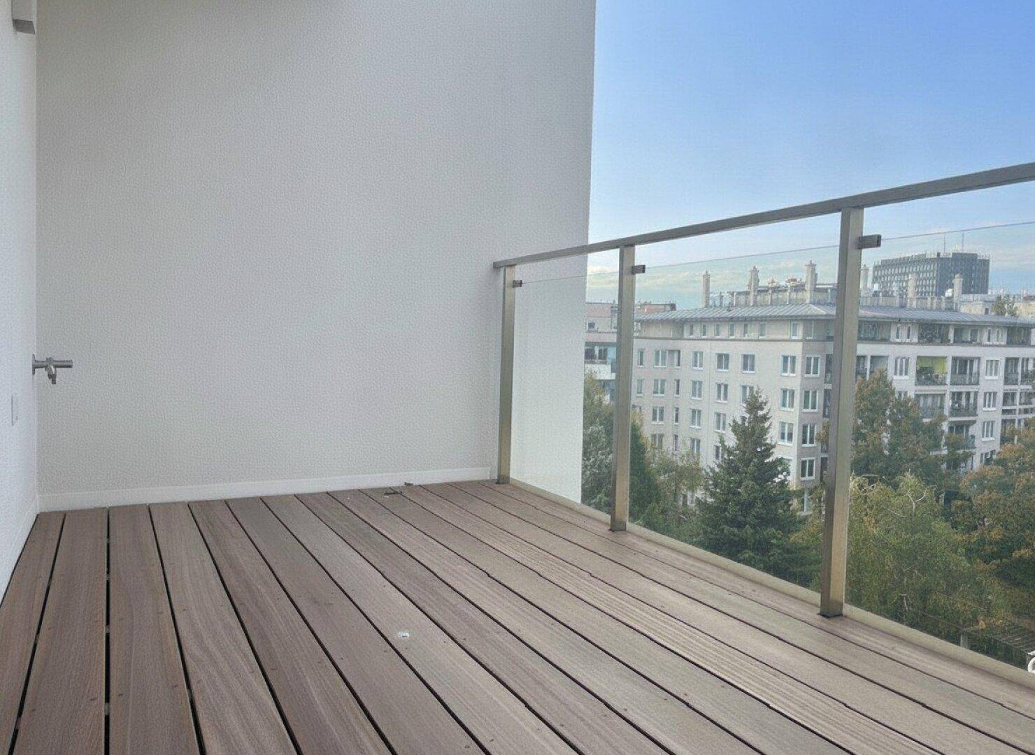Moderner Balkon mit Holzbelag, Glasgeländer und Blick auf die Stadt sowie grüne Bäume.