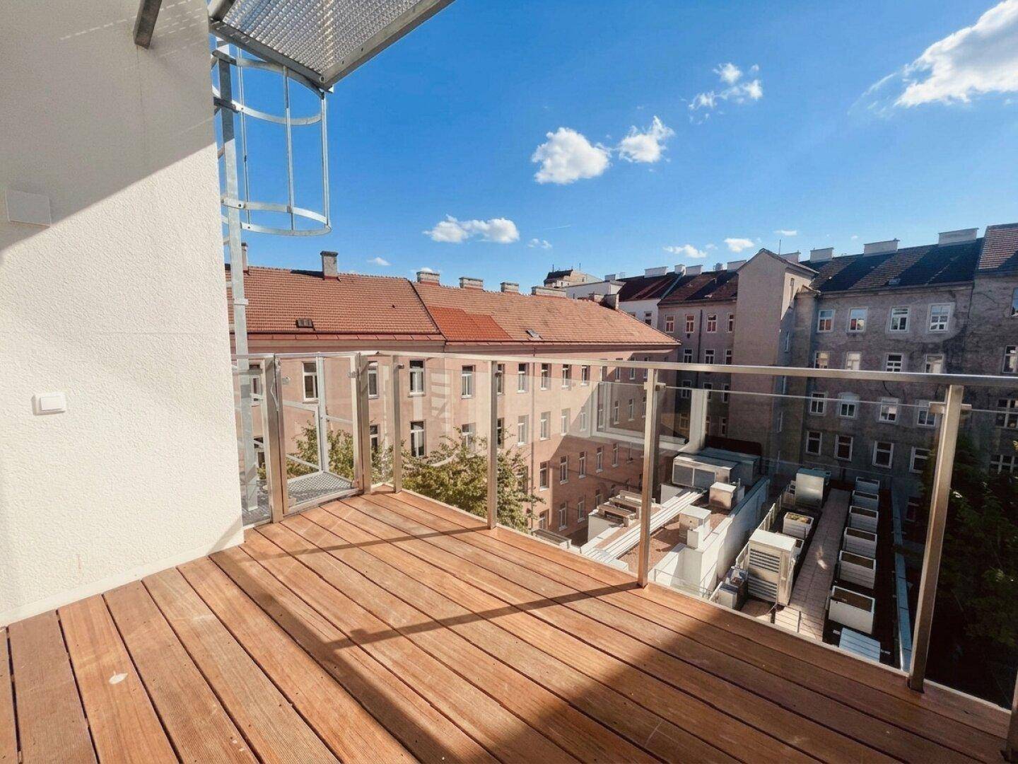 Sonniger Balkon mit Holzboden und Glasgeländer, bietet Blick auf die umliegenden Gebäude.