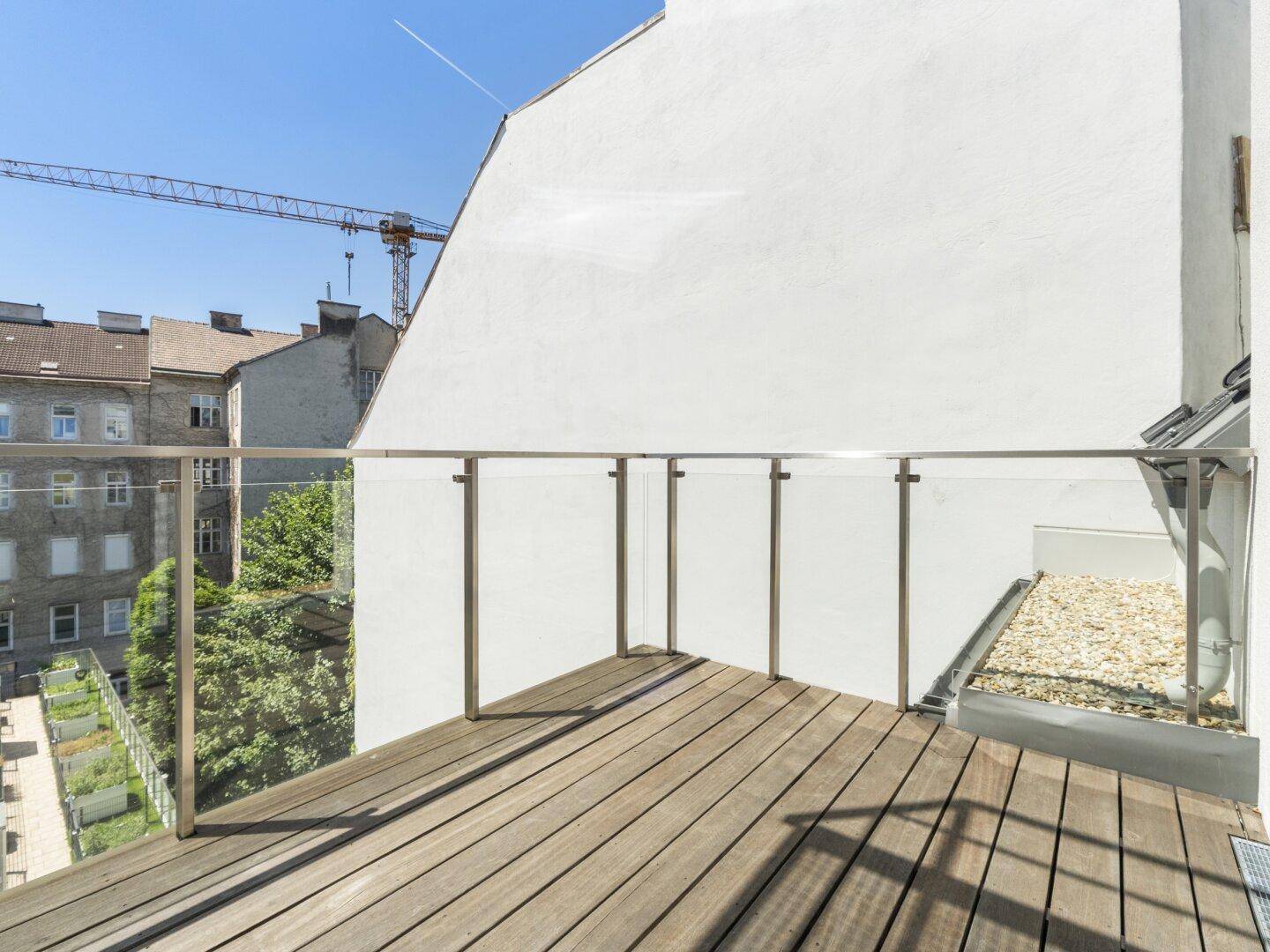 Geräumiger Balkon mit Holzboden und Glasgeländer, umgeben von städtischer Architektur.