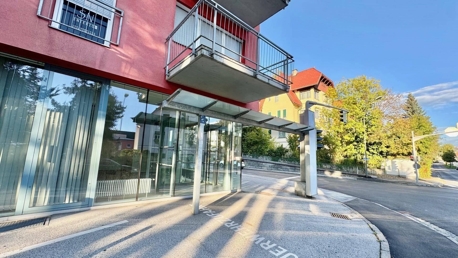 Gebäudeeingang mit Glasvordach und Balkon darüber, an einer sonnigen Straße gelegen.