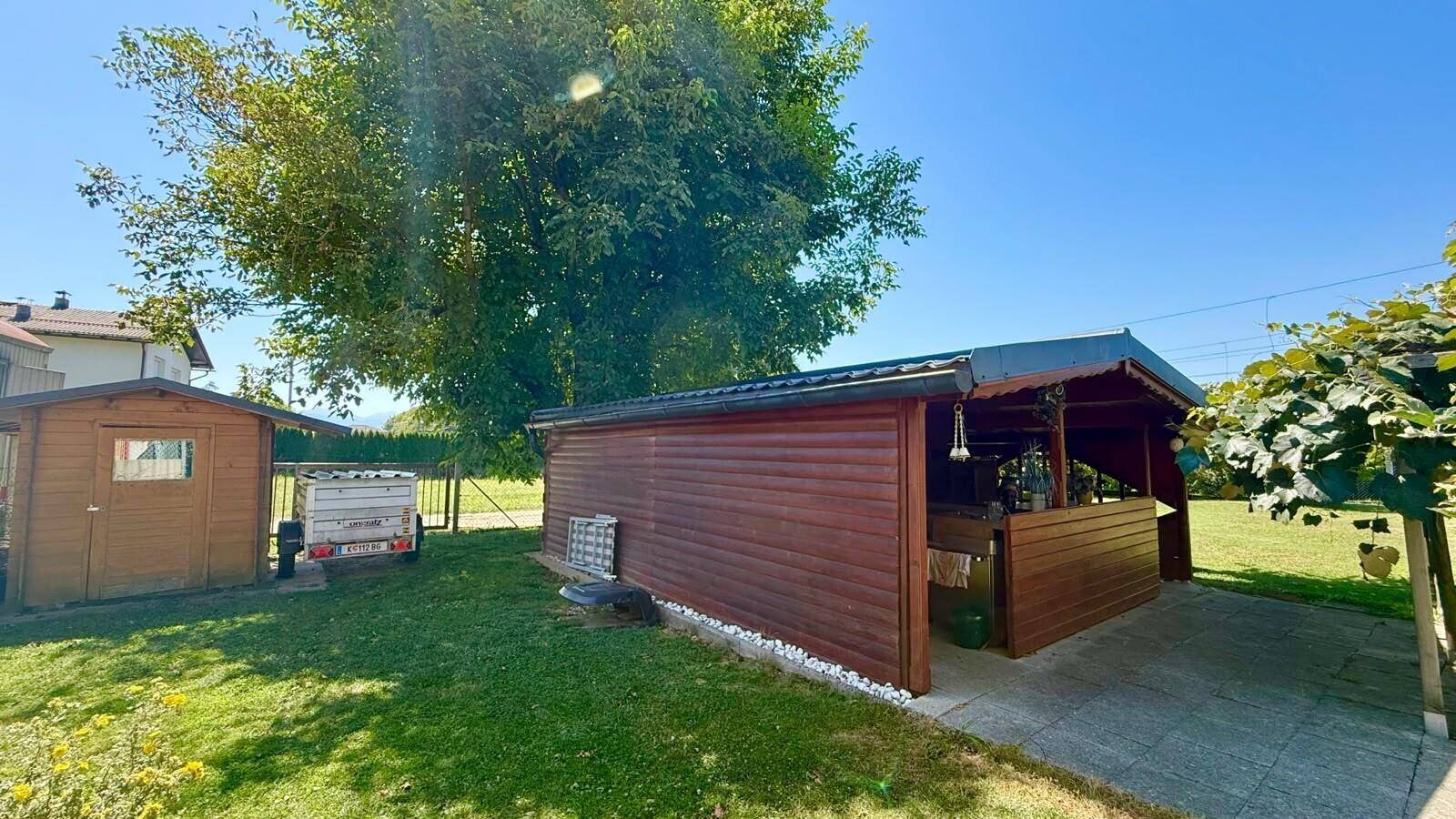 Gartenbereich mit einem Holzschuppen und einer überdachten Holzkonstruktion, umgeben von Rasen.