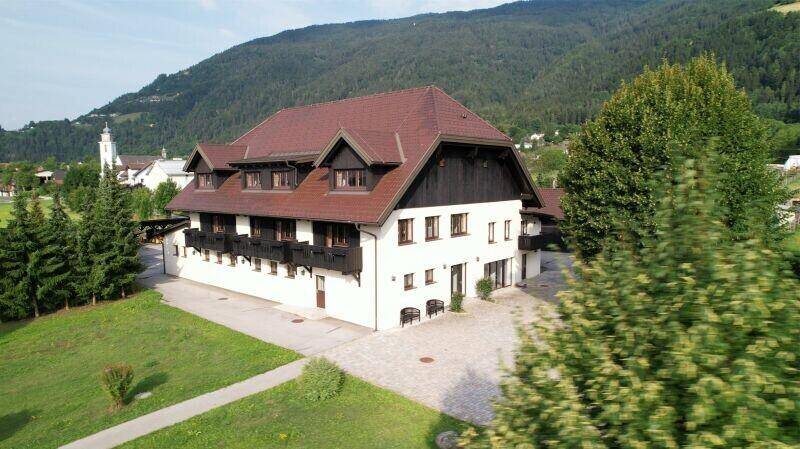 Traditionelles Gebäude mit braunem Ziegeldach und weißer Fassade, umgeben von grüner Landschaft und Bergen.