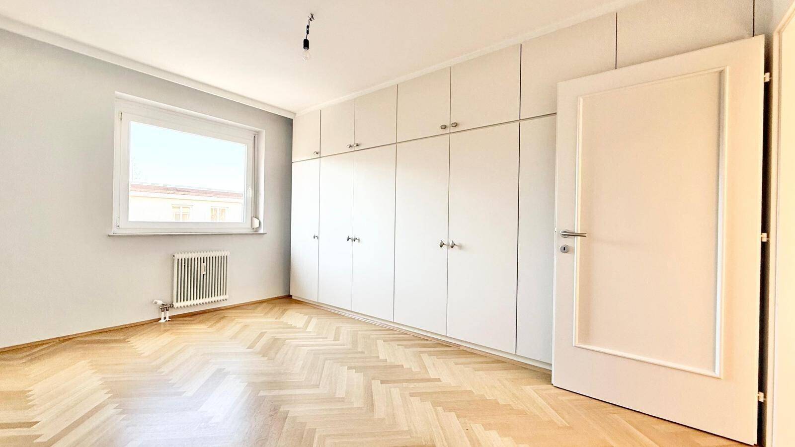 Helles Schlafzimmer mit großem Fenster, Einbauschränken und Fischgrät-Parkett.