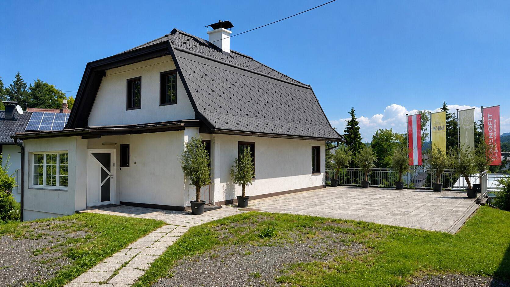 Haus mit gepflastertem Vorplatz und Grünflächen, umgeben von Bäumen und Pflanzen.