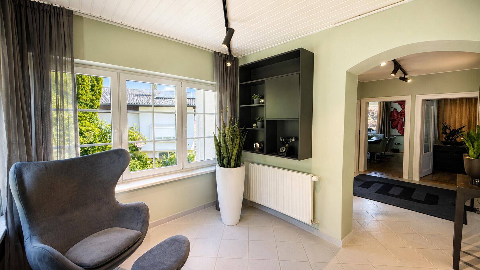 Heller Wohnbereich mit grün gestrichenen Wänden, Fliesenboden und einem modernen Sessel am Fenster.