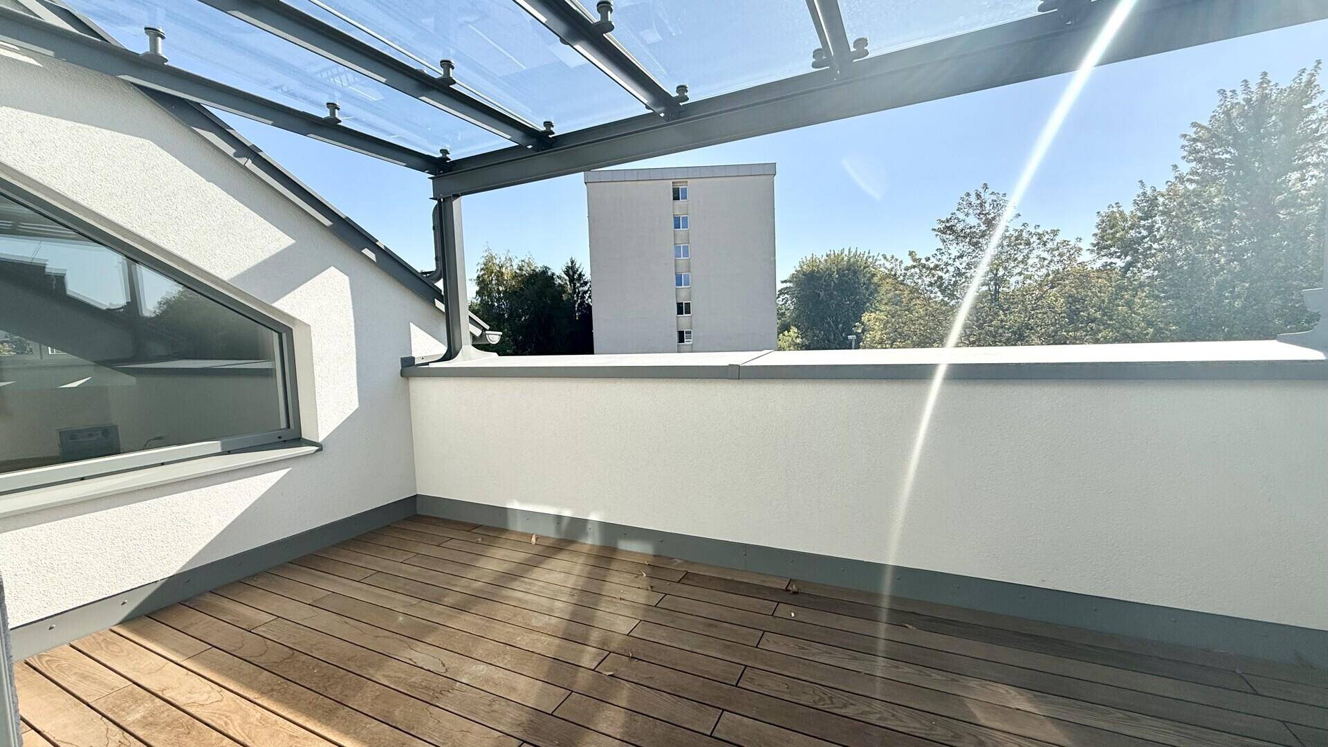 Sonniger Terrassenbereich mit Holzdeck, Glasdach und Blick auf die Umgebung.