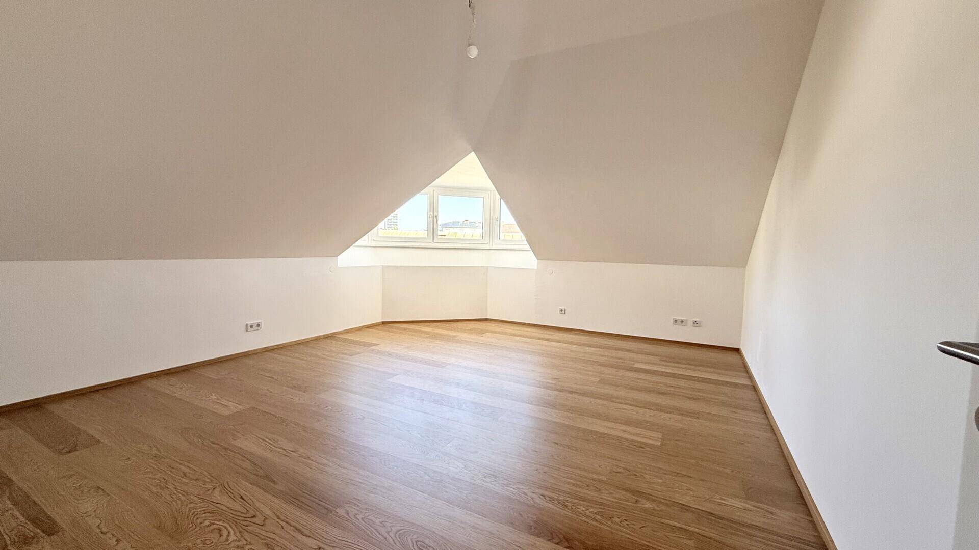 Heller Dachgeschossraum mit schrägen Wänden, Holzboden und einem dreieckigen Fenster.