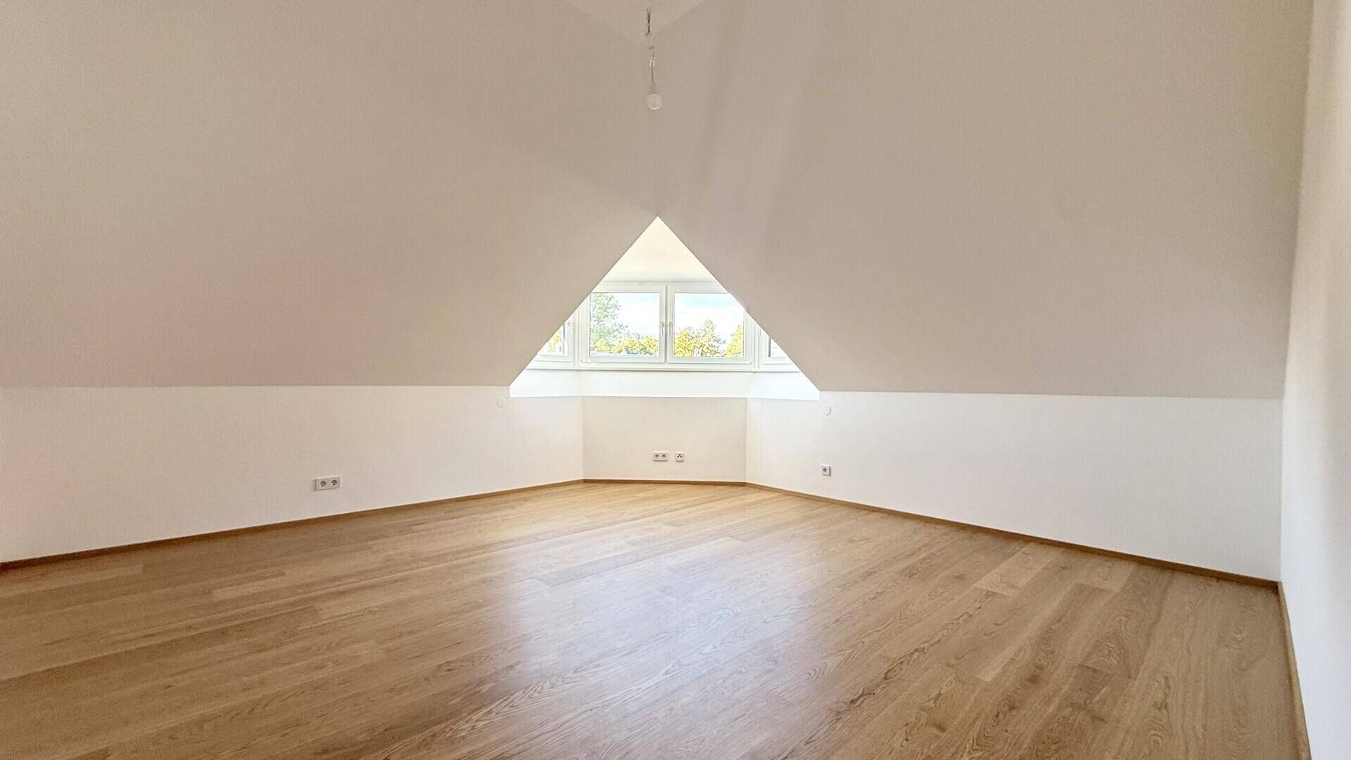 Lichtdurchfluteter Dachgeschossraum mit Holzboden und einem großen dreieckigen Fenster.