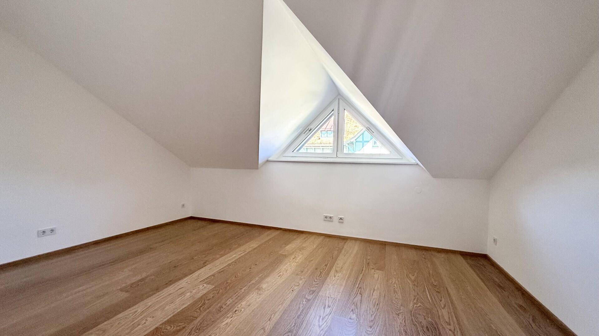 Geräumiger Raum im Dachgeschoss mit Holzboden und einem dreieckigen Fenster.