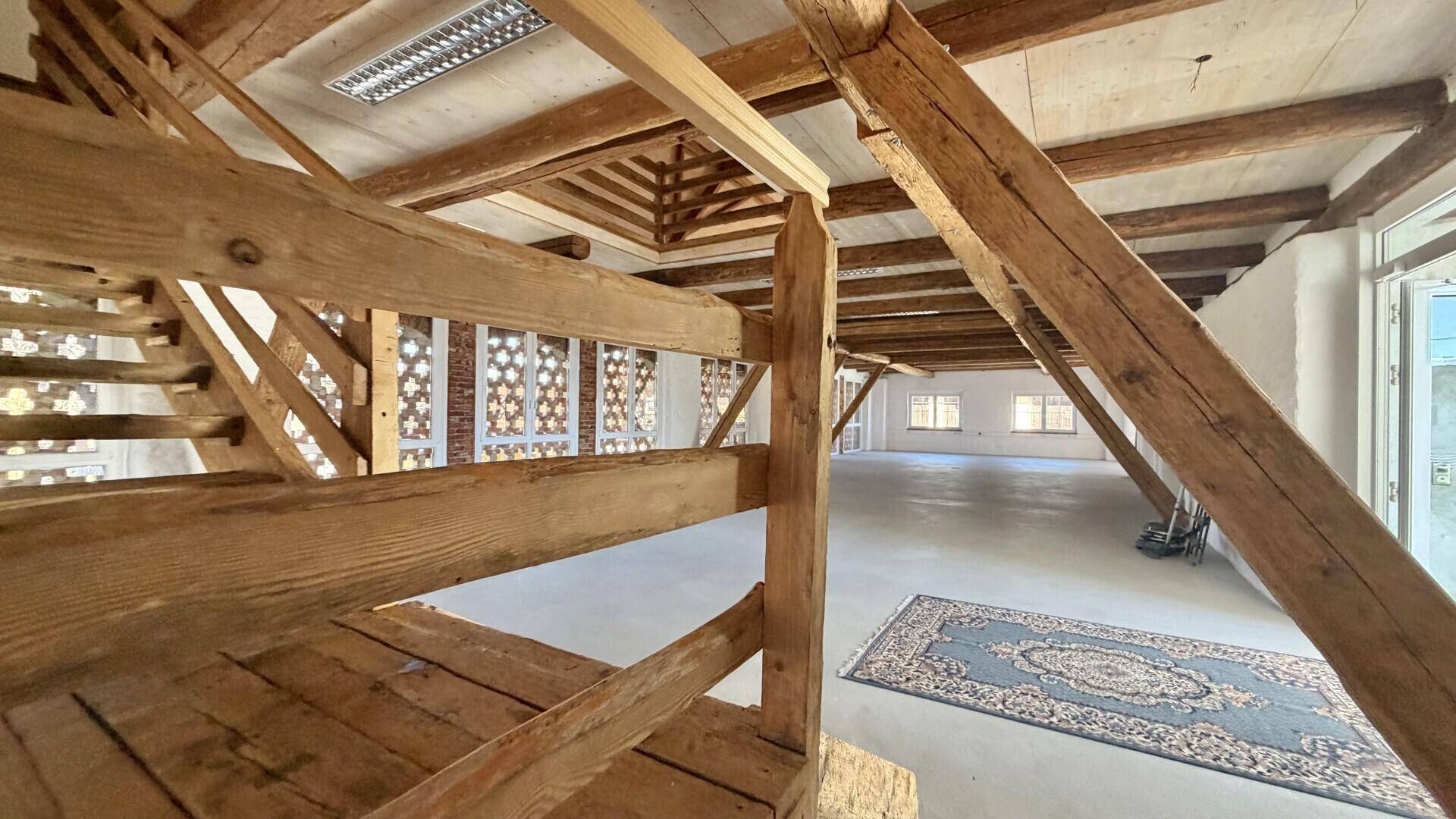 Offener Raum mit markanter Holztreppe und freiliegenden Balken, der vielseitige Nutzungsmöglichkeiten bietet.